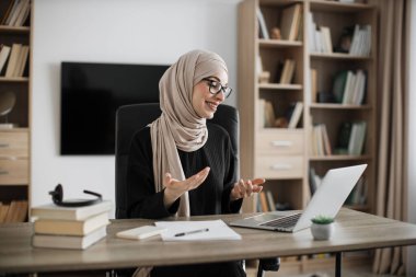 Smiling muslim businesswoman having video conversation while sitting at table and working on wireless laptop. Working process at office of young girl in headscarf.