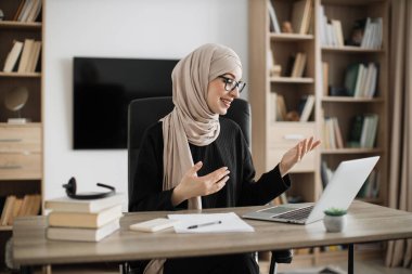 Smiling muslim businesswoman having video conversation while sitting at table and working on wireless laptop. Working process at office of young girl in headscarf.
