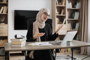 Smiling muslim businesswoman having video conversation while sitting at table and working on wireless laptop. Working process at office of young girl in headscarf.