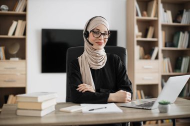 People, overwork, remote, deadline and office work concept. Confident smiling muslim woman in beige hijab sitting at desk with modern laptop and looking at camera at office.