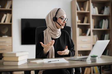 Young woman in headset having video call with partners, working process at office. Positive muslim business woman having conversation while sitting at table with laptop.