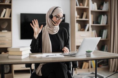 Young woman in headset having video call with partners, working process at office. Positive muslim business woman having conversation while sitting at table with laptop.