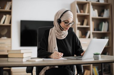 Attractive confident muslim business woman, office manager, wearing headset and hijab using laptop while making, writing financial report, using pen, on paper working indoors.