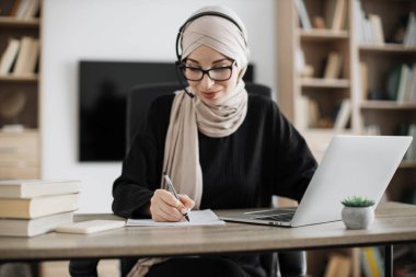 Attractive confident muslim business woman, office manager, wearing headset and hijab using laptop while making, writing financial report, using pen, on paper working indoors.