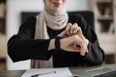 Happy muslim young businesswoman employee worker looking at smart watch sit at office desk with laptop, smartwatch digital modern technology apps for planning work time management concept