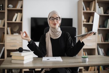 Muslim woman freelancer in eyeglasses sitting at table with closed eyes and relieving stress by meditation at workplace. Concept of relaxation and harmony, no stress free relief at work