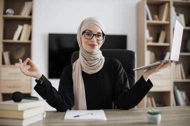 Muslim woman freelancer in eyeglasses sitting at table, relieving stress by meditation at workplace. Concept of relaxation and harmony, no stress free relief at work