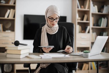 Rich muslim business woman working on laptop counting money cash, calculate earnings income profit at office workplace. Business people. Employment, occupation.