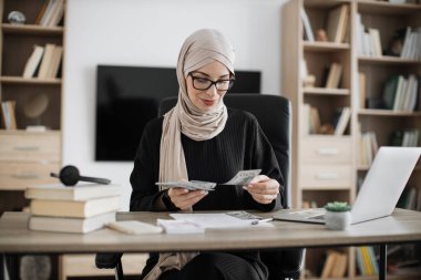Rich muslim business woman working on laptop counting money cash, calculate earnings income profit at office workplace. Business people. Employment, occupation.
