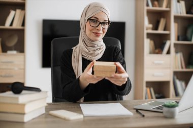 Smiling muslim woman in casual wear and hijab sitting at desk and holding gift box in hands. Famous female influencer opening festive present made by worldwide companies.
