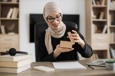 Excited muslim woman sitting at desk and opening parcel box. Female buyer checking ordered goods from delivery service. Concept of people and purchases.