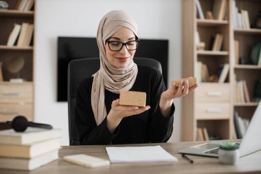 Excited muslim woman sitting at desk and opening parcel box. Female buyer checking ordered goods from delivery service. Concept of people and purchases.