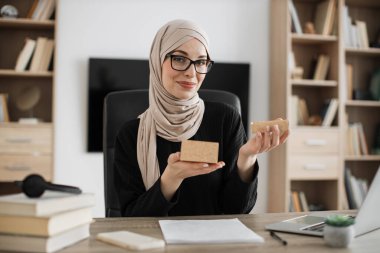 Excited muslim woman sitting at desk and opening parcel box. Female buyer checking ordered goods from delivery service. Concept of people and purchases.