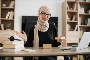 Attractive arabian woman with surprised facial expression looking inside paper box. Happy young lady in eyeglasses and hijab unpacking parcel.