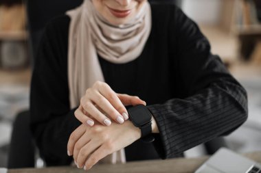 Cropped view of happy muslim young businesswoman employee worker looking at smart watch sit at office desk with laptop, smartwatch digital modern technology apps for planning work time.