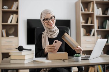 Muslim woman doing live stream while unpacking box with new smart phone. Female blogger sharing her feedback about modern gadget with her subscribers.