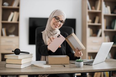 Muslim woman doing live stream while unpacking box with new smart phone. Female blogger sharing her feedback about modern gadget with her subscribers.