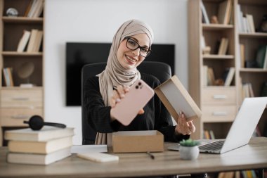 Muslim woman doing live stream while unpacking box with new smart phone. Female blogger sharing her feedback about modern gadget with her subscribers.