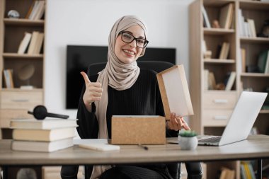 Excited muslim woman sitting at desk and opening parcel box. Female buyer checking ordered goods from delivery service showing thumb up. Concept of people and purchases.