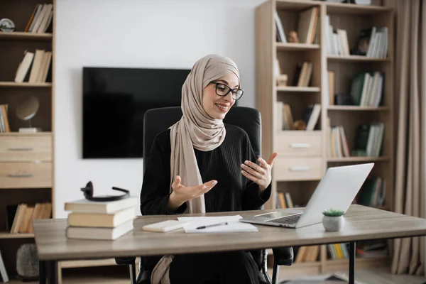 Smiling muslim businesswoman having video conversation while sitting at table and working on wireless laptop. Working process at office of young girl in headscarf.