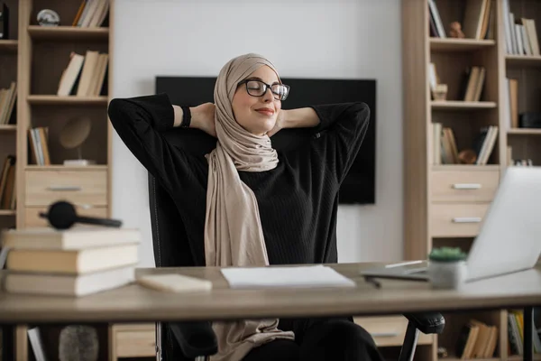 Gorgeous muslim woman relaxing by stretching her body while sitting in front her computer laptop at the wooden working desk over comfortable office or living room as background.