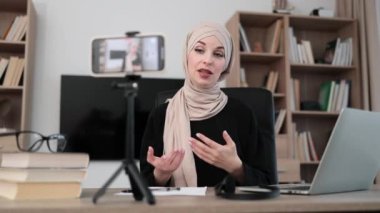 Charming muslim woman in hijab talking with subscribers while recording video on modern phone on tripod. Female blogger sitting at office and doing live stream.