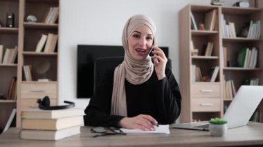 Muslim business woman having mobile conversation while sitting at table and working on wireless laptop. Working process at office of young arab girl in hijab.