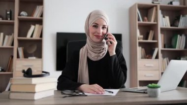 Muslim business woman having mobile conversation while sitting at table and working on wireless laptop showing sign ok. Working process at office of young arab girl in hijab.