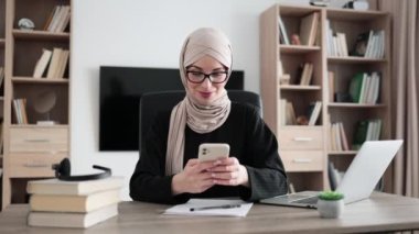 Beautiful young concentrated hardworking muslim businesswoman, it specialist or manager in hijab typing message on modern smartphone and working on laptop computer in office.