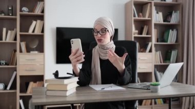 Portrait of smiling muslim businesswoman having mobile video conversation while sitting at table and working on wireless laptop. Working process at office of young girl in hijab.