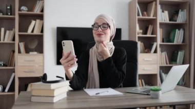 Young concentrated hardworking muslim businesswoman, it specialist or manager in hijab making selfie and showing air kiss using smart phone and working on laptop computer at office.