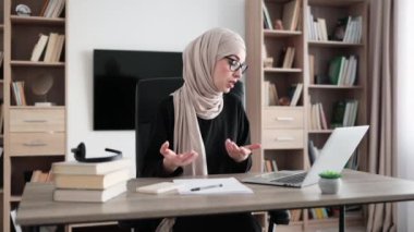 Smiling muslim businesswoman having video conversation while sitting at table and working on wireless laptop. Working process at office of young girl in headscarf.