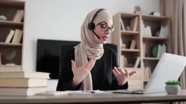 Young woman in headset having video call with partners, working process at office. Positive muslim business woman having conversation while sitting at table with laptop.