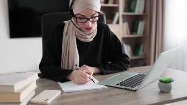 Attractive confident muslim business woman, office manager, wearing headset and hijab using laptop while making, writing financial report, using pen, on paper working indoors.