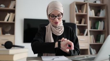 Young smiling happy successful employee muslim business woman in hijab looking at smartwatch time sit work at workplace wooden desk with laptop at office indoors. Achievement career concept.