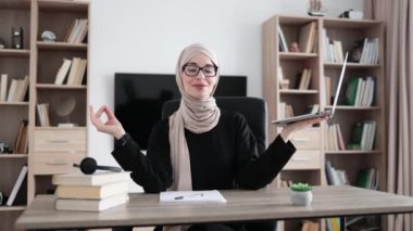 Muslim woman freelancer in eyeglasses sitting at table with closed eyes and relieving stress by meditation at workplace. Concept of relaxation and harmony, no stress free relief at work
