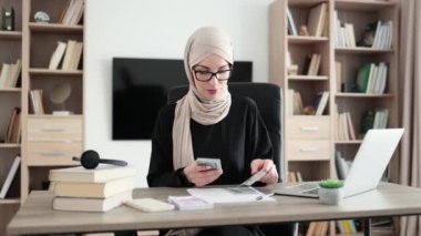 Rich muslim business woman working on laptop counting money cash, calculate earnings income profit at office workplace. Business people. Employment, occupation.