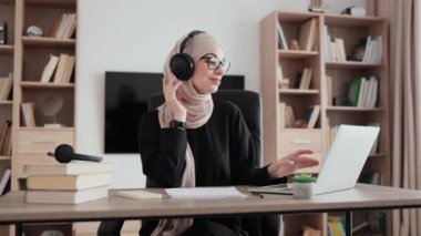 Portrait of a beautiful young muslim woman dressed in hijab, listening music with headphones and dancing. Relaxation during work. Break after study. Remote, freelance concept.