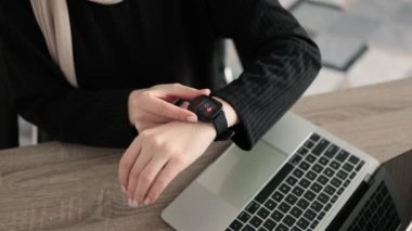 Cropped view of muslim female hands, putting finger on smartwatch, measuring heartbeat rate, while sitting at wooden table at office.