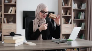 First person view of muslim female blogger opening gift box while sitting at home. Positive young woman doing live stream during unpacking process.