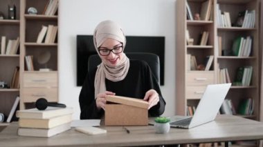 Excited muslim woman sitting at desk and opening parcel box. Female buyer checking ordered goods from delivery service. Concept of people and purchases.