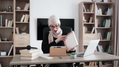 Excited muslim woman sitting at desk and opening parcel box. Female buyer checking ordered goods from delivery service. Concept of people and purchases.