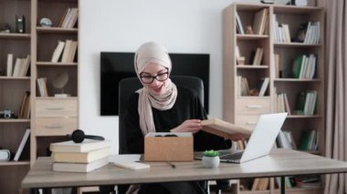 Excited muslim woman sitting at desk and opening parcel box. Female buyer checking ordered goods from delivery service showing thumb up. Concept of people and purchases.