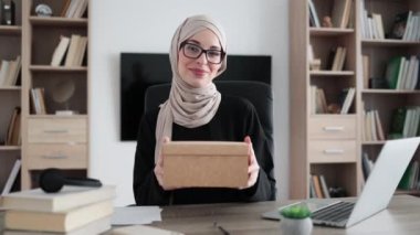 First person view of muslim female blogger opening gift box while sitting at home. Positive young woman doing live stream during unpacking process.