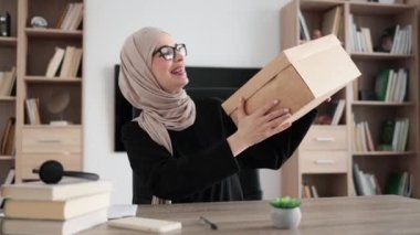 Excited muslim woman sitting at desk and opening parcel box. Female buyer checking ordered goods from delivery service. Concept of people and purchases.