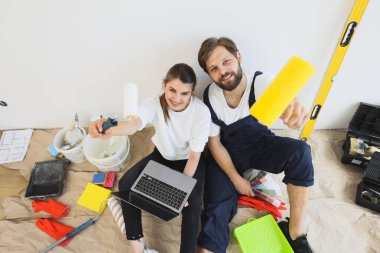 Joyful young wife and husband family couple sit on floor in new apartment during renovation process choosing new wall color and surfing internet on laptop choosing room design