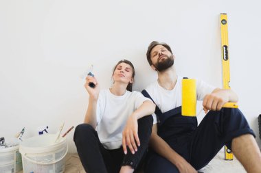 Tired young attractive caucasian couple sitting on the floor with paint brush and bucket with paints while doing repair at home.