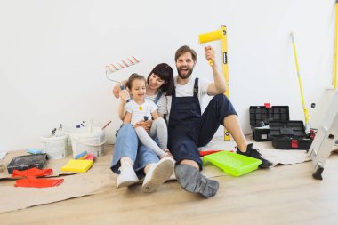 Young happy father, mother and their little cute girl daughter with wall paint rollers sitting on the floor during home renovation on white wall background.