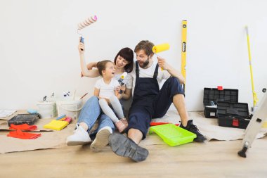 Young happy father, mother and their little cute girl daughter with wall paint rollers sitting on the floor during home renovation on white wall background.