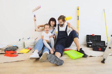 Cheerful caucasian family of father, mother and cute little girl daughter sitting on floor in white light room background with paint rollers in their hands.